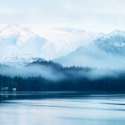 Blue waters of an Alaskan lake reflecting surrounding trees and distant mountains beneath a layer of mist