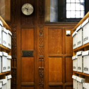 Narrow archive storage aisle in Oxford, UK, lined with shelves of labeled archive boxes, with a locked door and wall clock at the far end