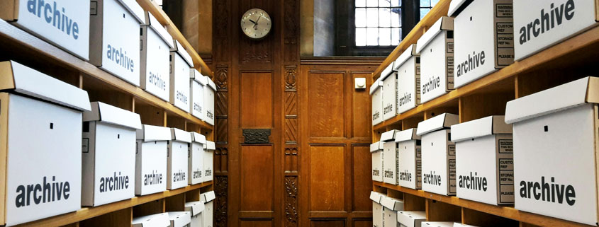 Archive-Storage-Room-With-Rows-of-Document-Boxes-Oxford-UK Narrow archive storage aisle in Oxford, UK, lined with shelves of labeled archive boxes, with a locked door and wall clock at the far end