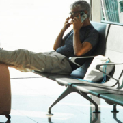 Man sitting in an airport waiting area, resting his legs on his luggage while talking on the phone, with his suit jacket hanging from the suitcase handle