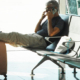 Man sitting in an airport waiting area, resting his legs on his luggage while talking on the phone, with his suit jacket hanging from the suitcase handle