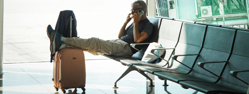 Business-Traveler-Relaxing-With-Luggage-at-Airport-During-Layover Man sitting in an airport waiting area, resting his legs on his luggage while talking on the phone, with his suit jacket hanging from the suitcase handle