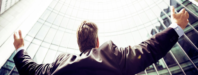 Businessman-Looking-Up-With-Raised-Arms-Outside-Office-Building Businessman standing outside a modern office building with both arms raised while looking upward