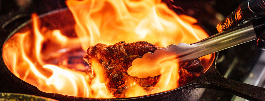 Chef using kitchen tongs to turn meat in a frying pan with controlled flames rising above the food and heat from the stove below