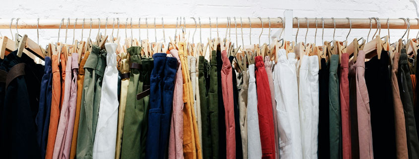 Colorful assortment of men's and women's clothing displayed on hangers along a wooden rail mounted against a white wall