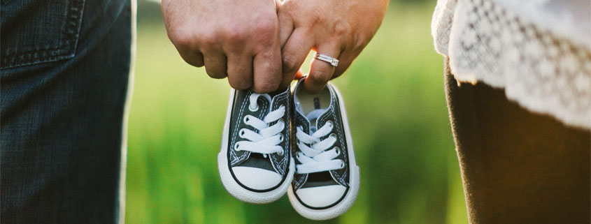 Couple-Holding-Hands-With-Tiny-Baby-Shoes-and-Wedding-Ring Couple holding hands while each holds a tiny baby shoe, with a wedding ring visible on the woman's hand