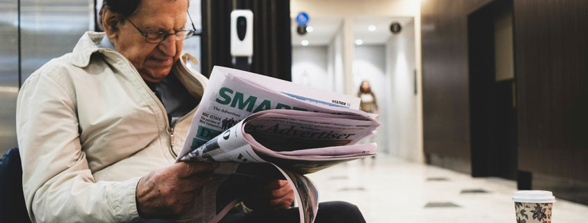 Elderly man wearing glasses and reading a newspaper with a serious, thoughtful expression