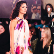 Elegantly dressed woman walking in a long gown while carrying a handbag as spectators watch at a fashion event