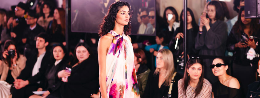 Elegantly dressed woman walking in a long gown while carrying a handbag as spectators watch at a fashion event