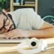 Exhausted man sleeping with his head resting on a closed laptop, surrounded by notebooks, headphones, a phone, and a cup of coffee