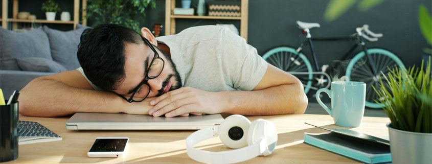Exhausted man sleeping with his head resting on a closed laptop, surrounded by notebooks, headphones, a phone, and a cup of coffee