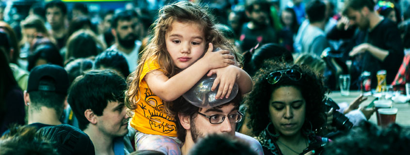 Man carrying a young girl on his shoulders amid a densely packed crowd of people in a busy public area
