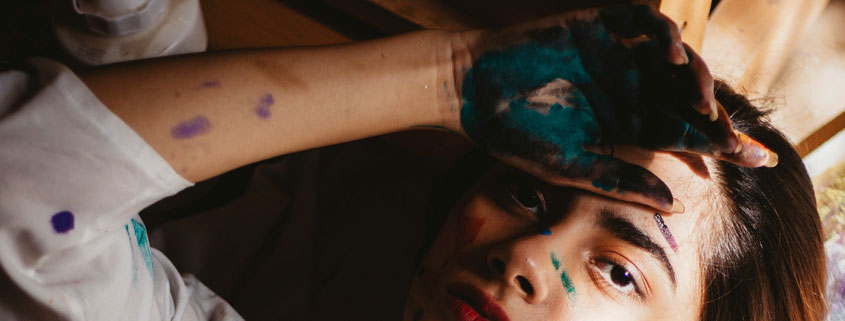 Female-Artist-Lying-Down-With-Paint-Covered-Hands-and-Brush Female artist lying down with paint on her shirt, hands, and fingers while holding a paintbrush and resting one hand on her forehead