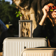 Woman traveling with two suitcases, resting one hand on her luggage while holding a camera to photograph something outside the frame
