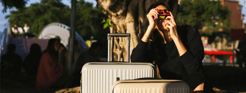 Woman traveling with two suitcases, resting one hand on her luggage while holding a camera to photograph something outside the frame