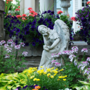 Flower garden featuring an angel statue surrounded by plants near a porch with classical columns