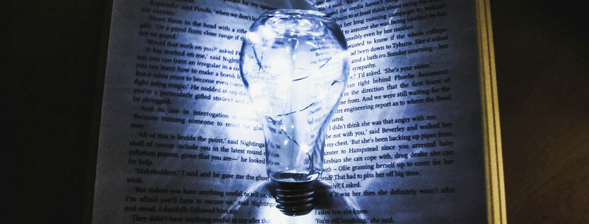 Glowing-Light-Bulb-Illuminating-Open-Book-in-Darkness Glowing light bulb positioned at the center of an open book, illuminating its pages against a dark background