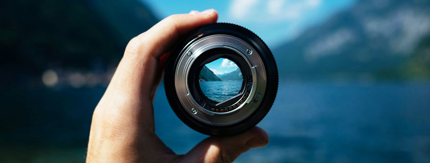 Hand-Holding-Camera-Lens-Framing-Mountain-Lake-Landscape Hand holding a detached camera lens that frames a mountain lake surrounded by towering mountains