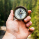 Hand holding a navigation compass toward a distant forest while searching for direction through the natural landscape