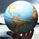 Wet hand holding a spherical globe representing Earth above the water, with a lake visible in the background