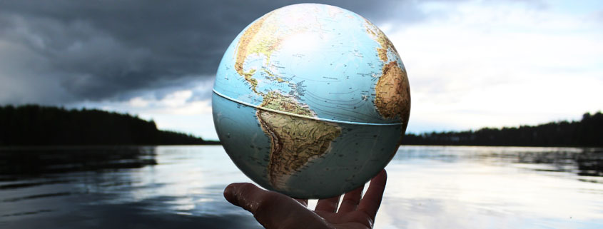 Hand-Holding-World-Globe-Above-Water-With-Lake-in-Background Wet hand holding a spherical globe representing Earth above the water, with a lake visible in the background