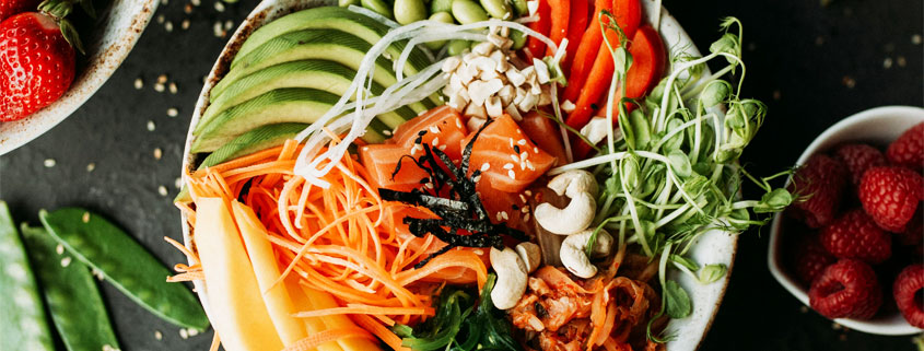 Colorful assortment of healthy foods featuring strawberries, avocado, blueberries, mango, carrots, nuts, and sushi arranged on a plate