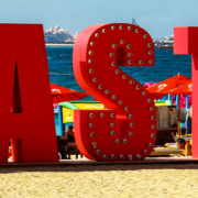 Large sign displaying the word Taste on a sandy beach with boats and cruise ships visible across the distant water