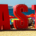 Large sign displaying the word Taste on a sandy beach with boats and cruise ships visible across the distant water
