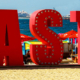 Large sign displaying the word Taste on a sandy beach with boats and cruise ships visible across the distant water