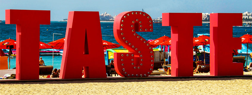 Large sign displaying the word Taste on a sandy beach with boats and cruise ships visible across the distant water