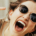 Woman wearing sunglasses and laughing with her mouth wide open, showing white teeth while making playful gestures with her hands and fingers