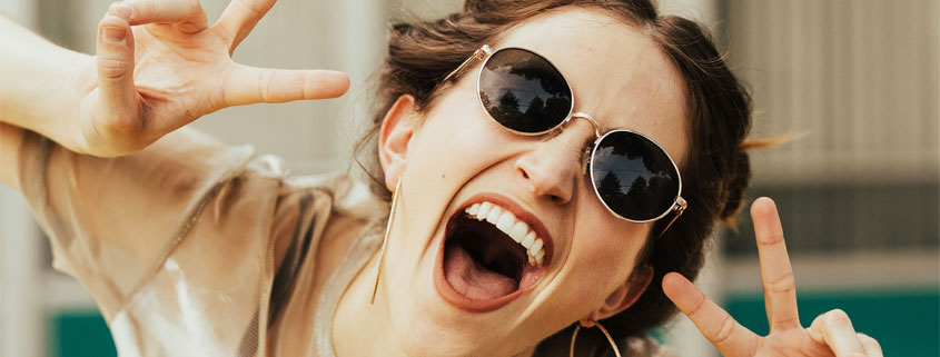 Laughing-Woman-With-Sunglasses-White-Teeth-and-Hand-Gestures Woman wearing sunglasses and laughing with her mouth wide open, showing white teeth while making playful gestures with her hands and fingers