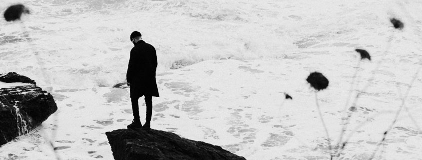 Lone-Man-Standing-on-Rock-Cliff-Above-Powerful-Ocean-Waves Lone man standing near the edge of a rocky coastal cliff overlooking powerful ocean waves crashing against the rocks below