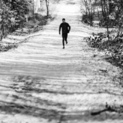 Lone man running uphill along a snow-covered forest trail surrounded by tall trees on both sides