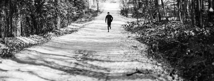 Lone-Runner-on-Snow-Covered-Forest-Trail-Up-Hill Lone man running uphill along a snow-covered forest trail surrounded by tall trees on both sides