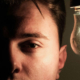 Man looking straight ahead with one side of his face illuminated by a glowing light bulb while the opposite side remains in darkness