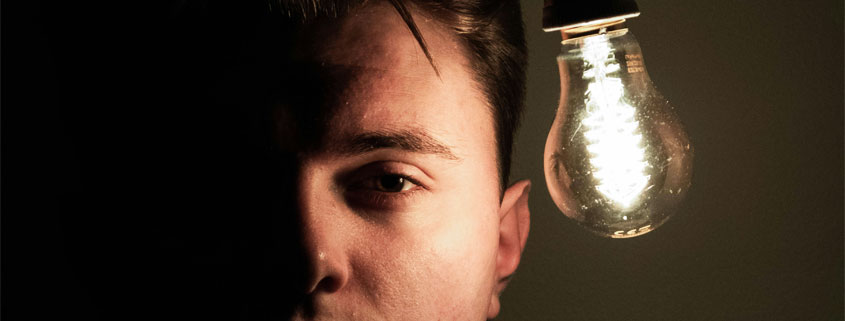 Man-Face-Partially-Illuminated-by-Glowing-Light-Bulb Man looking straight ahead with one side of his face illuminated by a glowing light bulb while the opposite side remains in darkness