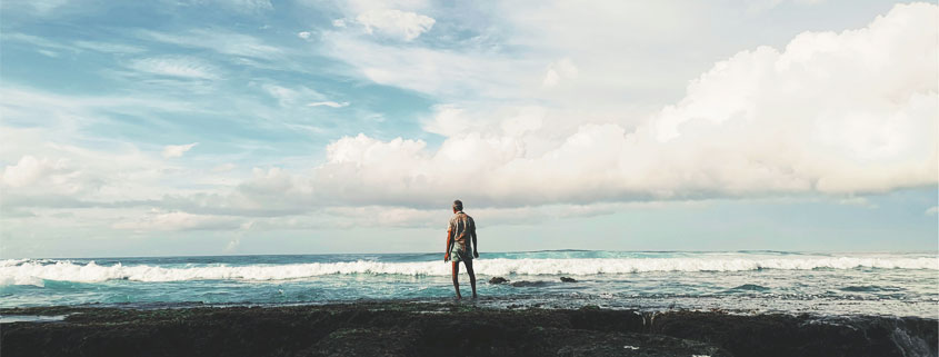 Man-Standing-Alone-on-Shore-Watching-Ocean-Waves Man standing alone on the shoreline, looking out toward the sea as large waves roll across the water