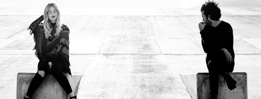 Man-and-Woman-Sitting-Apart-on-Concrete-Ramps-in-Quiet-Moment Man and woman sitting separately on two concrete ramps, with the man looking behind him and the woman closing her eyes while touching her hair
