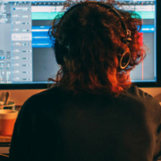 Rear view of a musician wearing headphones and playing guitar in front of two computer monitors surrounded by music equipment and a keyboard