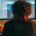 Rear view of a musician wearing headphones and playing guitar in front of two computer monitors surrounded by music equipment and a keyboard