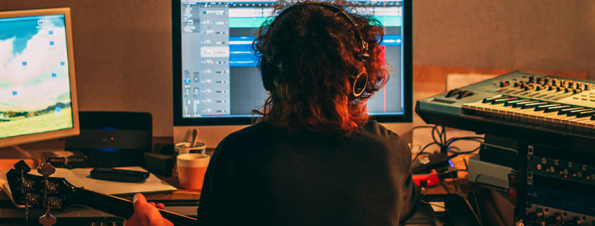 Musician-With-Guitar-Headphones-and-Dual-Monitor-Music-Setup Rear view of a musician wearing headphones and playing guitar in front of two computer monitors surrounded by music equipment and a keyboard