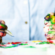 Two hands covered in colorful paint holding a pencil and paintbrush above a paint-splattered work surface