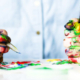 Two hands covered in colorful paint holding a pencil and paintbrush above a paint-splattered work surface