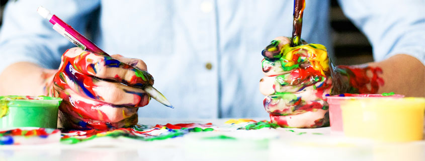Two hands covered in colorful paint holding a pencil and paintbrush above a paint-splattered work surface