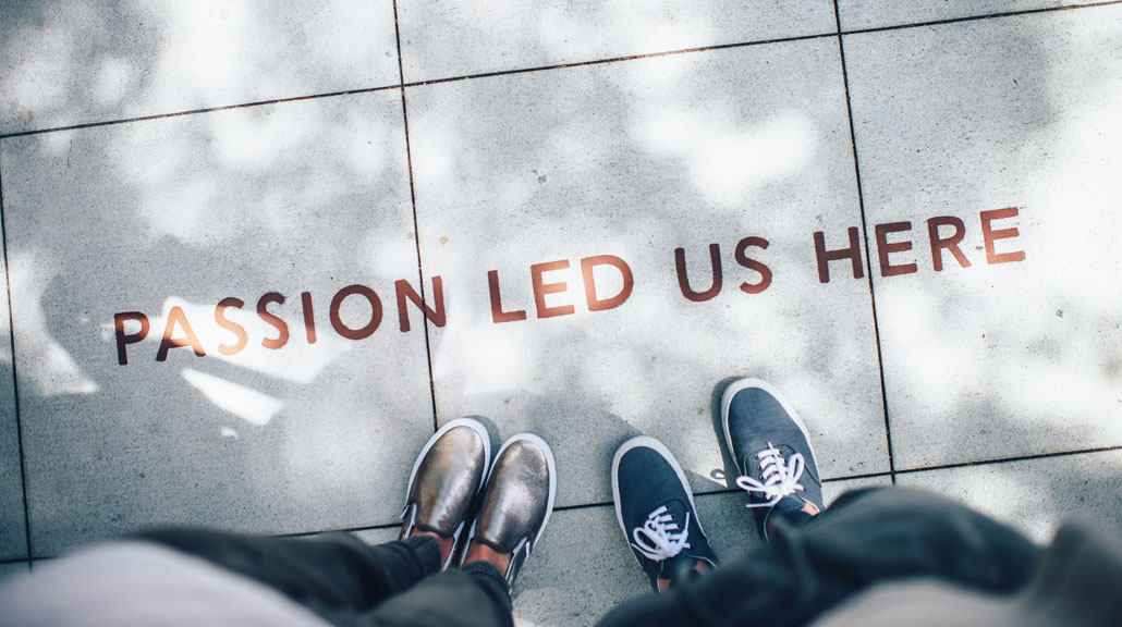 Passion-Led-Us-Here-Inspiring-About-Us-Message Two people standing side by side with their shoes visible beside a floor sign displaying the inspirational message “Passion Led Us Here”