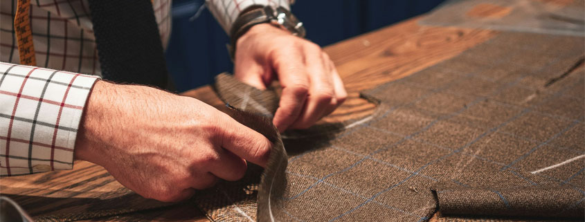 Professional-Tailor-Cutting-and-Sewing-Fabric-at-Worktable Professional tailor working with fabric at a cutting table, wearing a measuring tape around his neck while preparing and sewing a garment