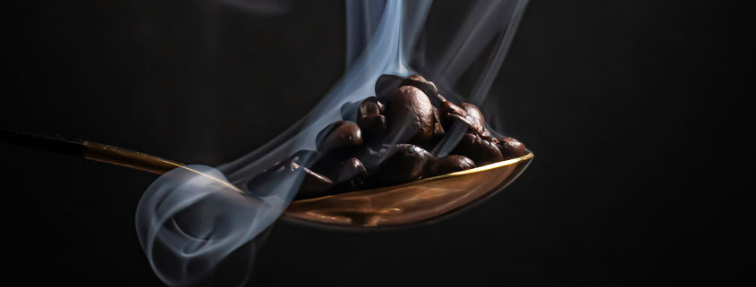 Roasted-Coffee-Beans-on-Spoon-With-Rising-Aromatic-Steam Spoon filled with roasted coffee beans surrounded by delicate wisps of white smoke or steam rising from the beans