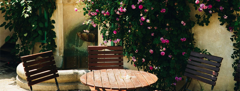 Rose-Covered-Garden-Wall-With-Outdoor-Table-Chairs-and-Fountain Small garden with an outdoor table and chairs, a wall covered in climbing roses, and a water fountain in the background