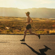 Shirtless male runner wearing athletic shorts, running shoes, a hat, and sunglasses while jogging along a road in Arizona